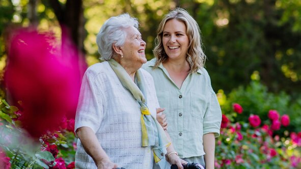 Senior woman walking arm in arm with a young Herewith Helper through a blooming rose garden outdoors.