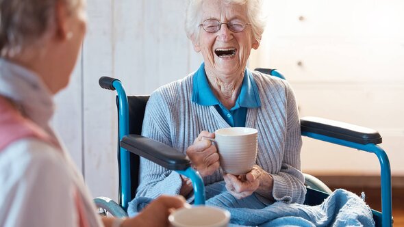 A senior woman in a wheelchair bursts out laughing while holding a mug, enjoying a lighthearted moment with her Helper. The mood is bright and cheerful.