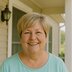 A woman in a light teal shirt stands on a porch smiling directly at the camera.