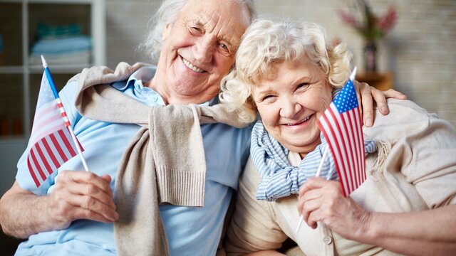 A senior couple sits closely together, smiling and holding small American flags. They appear patriotic, joyful, and proud of their country.