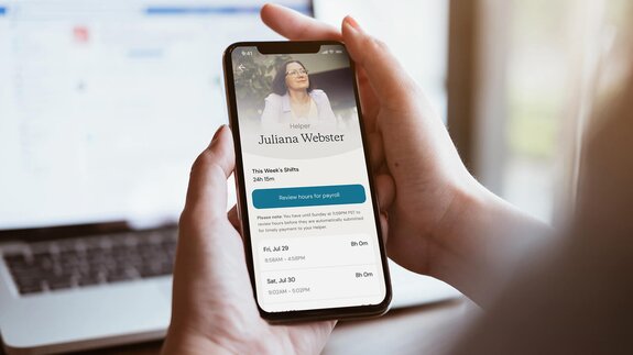 A person holds a phone showing a Helper's timesheet with hours worked and a button for payroll approval.