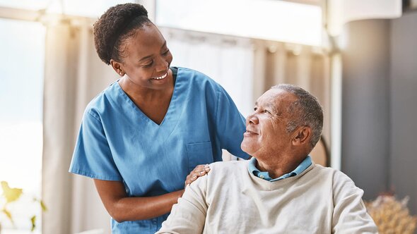 A Helper stands beside an older man indoors and rests a hand on his shoulder as they look at each other.