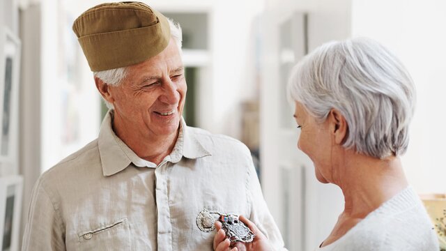 A senior man wearing a military-style cap smiles warmly as a Helper hands him a medal. They share a joyful moment, possibly celebrating his service or an achievement.