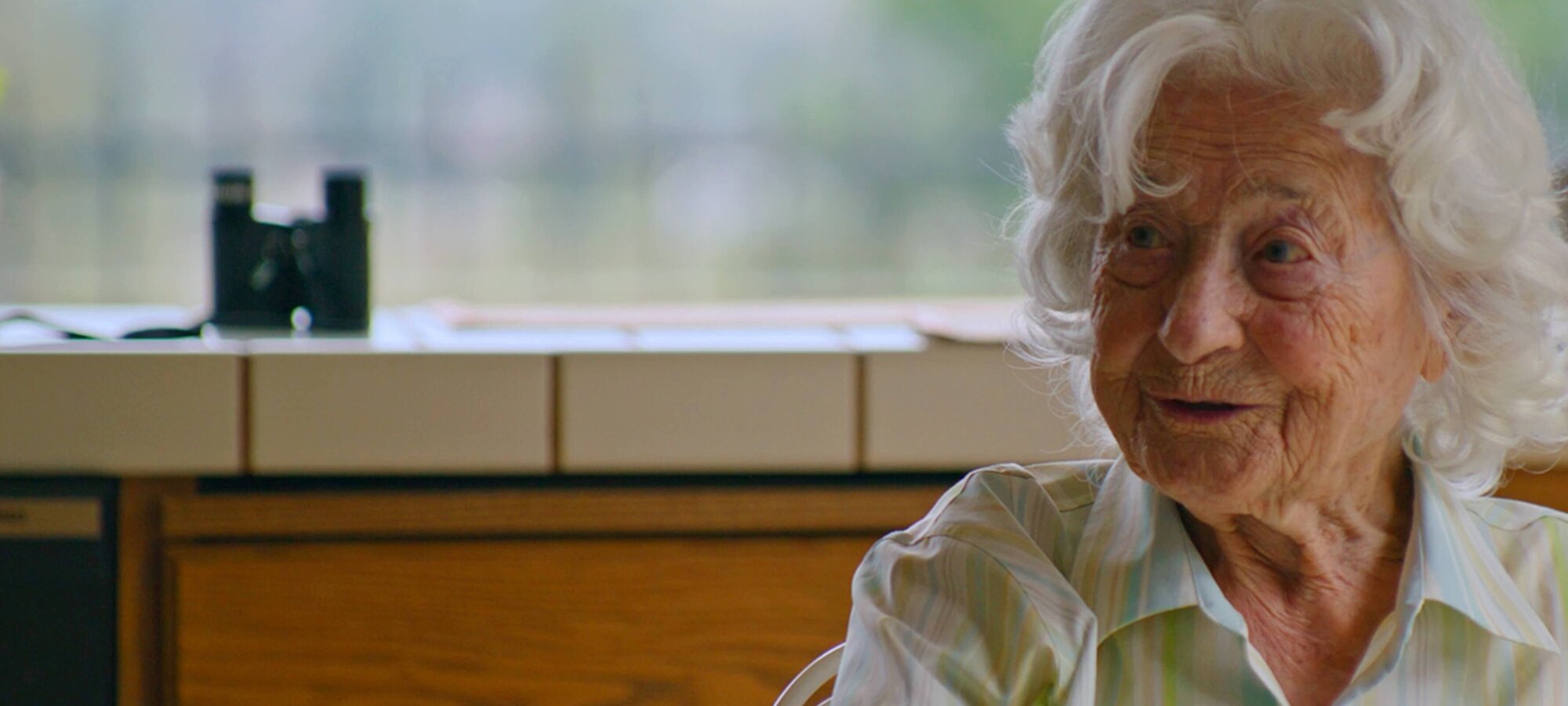 A senior woman with white hair sits at a kitchen table, gesturing with her hand as she speaks. She appears to be mid-conversation, possibly sharing a story or memory. The mood is thoughtful and engaged.
