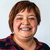 A woman in a plaid shirt smiles at the camera in front of a plain background.