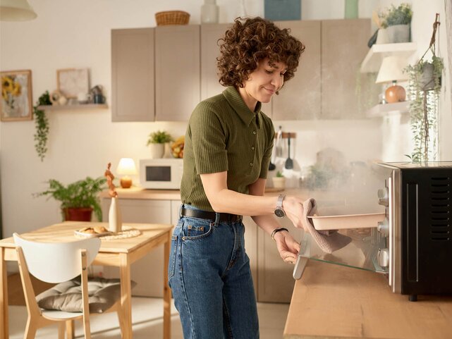 Woman in an olive shirt lifts a tray from the oven in a bright home kitchen, smoke is pouring out of the oven