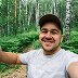 A man in a cap takes a selfie in a wooded area with green foliage and trees behind him.