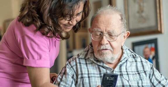A Helper shows a smartphone to a senior man in glasses who is looking at the screen while seated indoors.