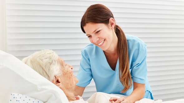 A Helper in scrubs leans in and smiles at an older woman lying in bed.