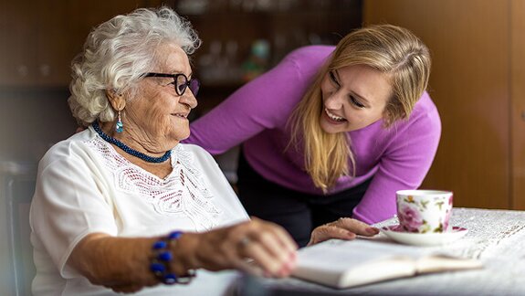 A Helper leans over a table to smile at an older woman who is looking at a book with a teacup nearby.