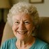 A senior woman with curly white hair smiling brightly while seated at home.