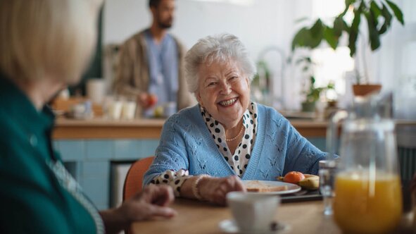 Happy senior woman laughing at the breakfast table while a Herewith Helper prepares a healthy morning meal.