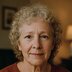 A woman with short curly hair looks at the camera with a neutral expression while sitting indoors.