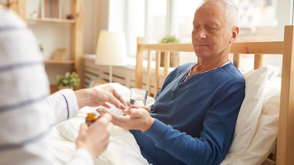 A Helper hands a pill and glass of water to a senior man sitting up in bed.