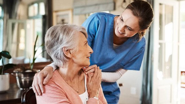 Herewith Helper in scrubs smiling and resting a reassuring hand on a senior woman's shoulder at home.