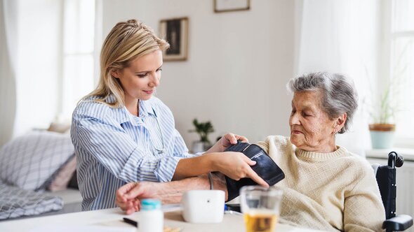 A Helper wraps a blood pressure cuff around the arm of an older woman in a wheelchair seated at a table.
