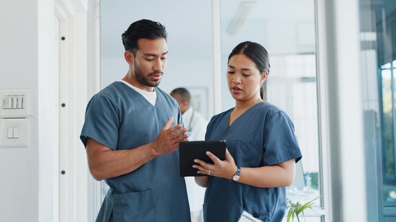 Two healthcare professionals in scrubs review information together on a tablet while standing in a medical facility hallway.