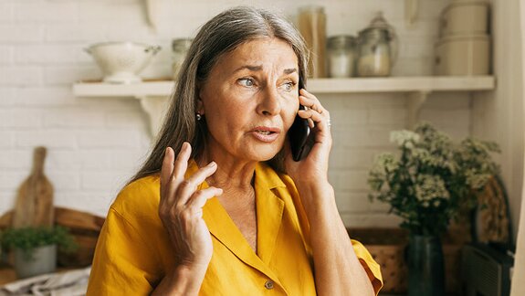 An older woman with long hair speaks on the phone while standing in a kitchen.