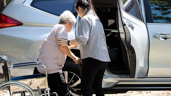 A Helper assists a senior woman using a cane and wheelchair to get out of a car, with sunlight highlighting the scene.