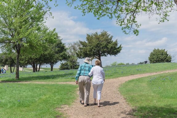 Two seniors walk in the park arm in arm. There's trees in the background and the weather is sunny with blue skies.
