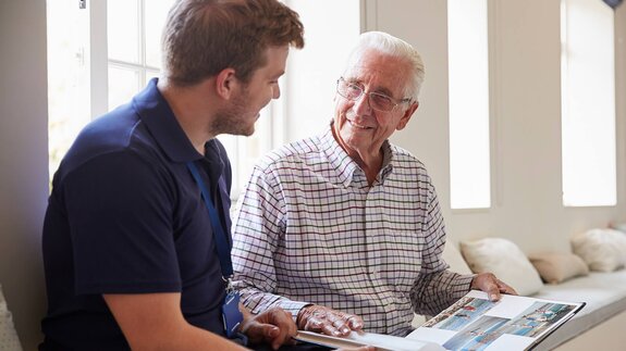 A Helper sits beside an older man as they talk and look through a photo album together.