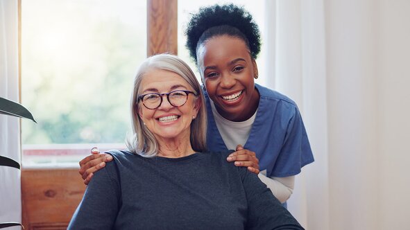 A Helper and a senior smile warmly at the camera, the senior is seated and wearing glasses with long silver hair. The caregiver has her hair pulled back in a bun and has blue scrubs on. They're inside a home