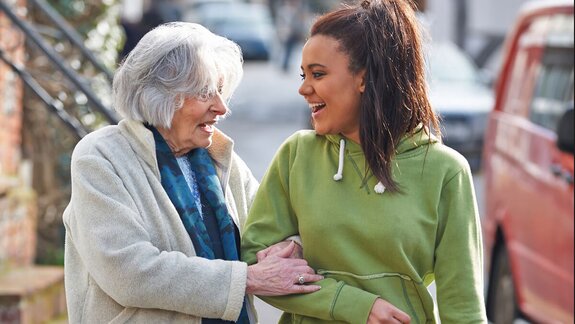 A senior and a younger woman laughing together while walking arm-in-arm outdoors, with cars and stairs in the background.