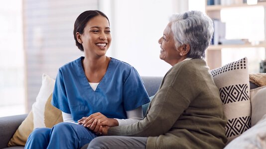 Herewith Helper in scrubs smiling and holding hands with a senior woman on the sofa during a companion care visit.