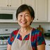 A woman wearing an apron stands and smiles in a kitchen.
