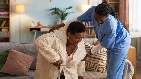 A Helper in blue scrubs supports an older woman with a cane as she stands up from her living room sofa.