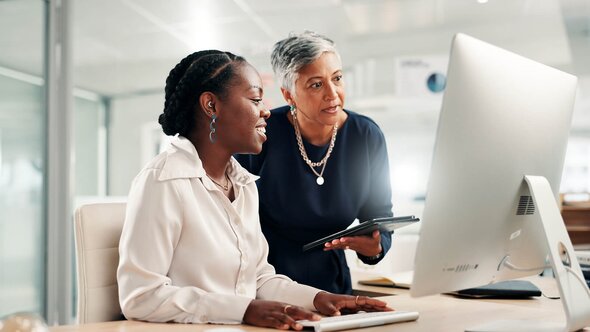 Two healthcare workers look at a computer screen while one holds a tablet and speaks.