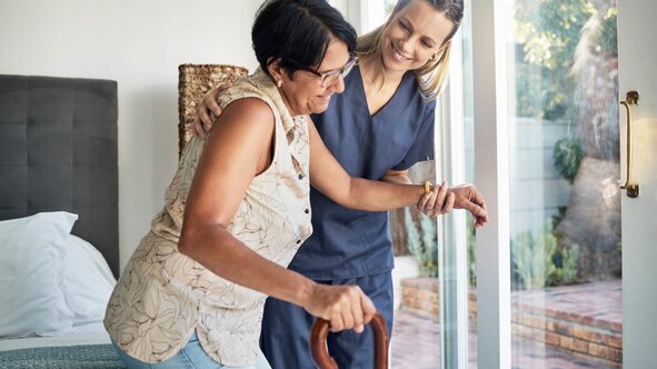 Herewith Helper in scrubs helping a senior woman stand up with her cane, providing mobility and fall-prevention support.