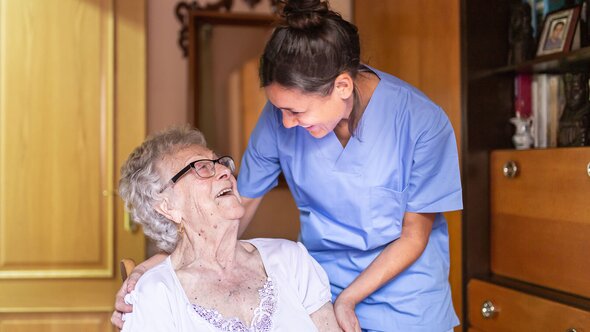 A Helper in scrubs smiles and speaks with an older woman seated in a chair in her home.