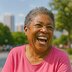A woman in a pink shirt laughs while sitting outside in a park with trees and city buildings in the background.