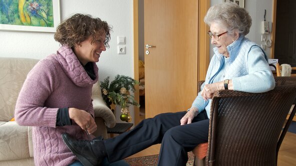 A Helper kneels to help an older woman put on her shoes as she sits in an armchair at home, both smiling.