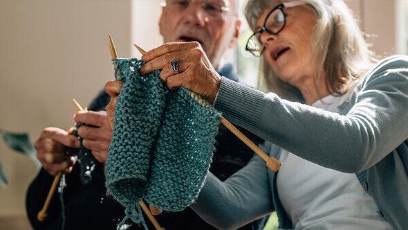 Two older adults sit closely together, knitting with blue yarn and wooden needles. The atmosphere is focused and warm, suggesting shared learning or companionship.