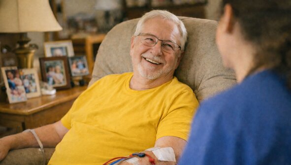 A man sits in his living room chair undergoing home hemodialysis treatment. He's supported by a Herewith Helper, they're smiling and talking. 