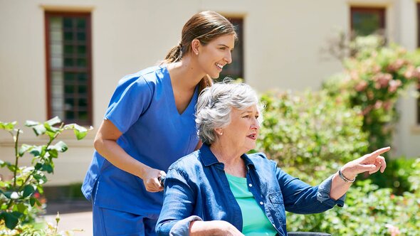 A Helper pushes an older woman in a wheelchair outside while she points toward something in the distance.