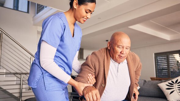 A Helper supports an older man as he rises from a chair in a senior home.