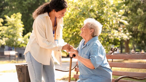 A Helper holds both hands of a senior woman who is sitting on a bench with a cane, and they are facing each other.