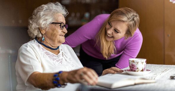 A Helper leans over a table and talks with a senior woman who is seated and smiling, with a teacup and open book in front of them.