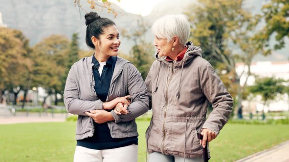 A Helper walks arm in arm with an older woman using a cane along a tree-lined park path.