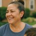 A woman smiles while standing outside near a residential building, with a child partially visible in the corner.