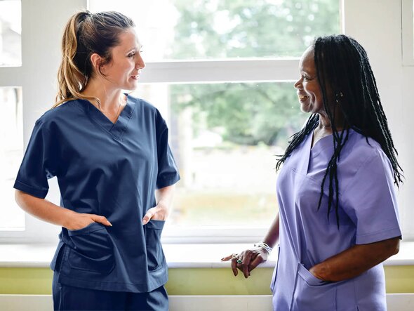 Two caregivers in scrubs stand and talk together near a window in a bright indoor space.