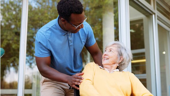 A Helper speaks with a senior woman seated in a wheelchair outside a building, and they are both smiling.