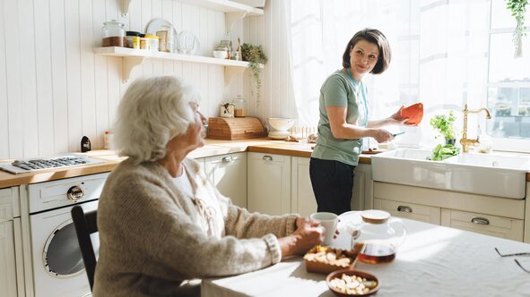 A Helper prepares a meal at the sink while an older woman chats with her from the kitchen table.