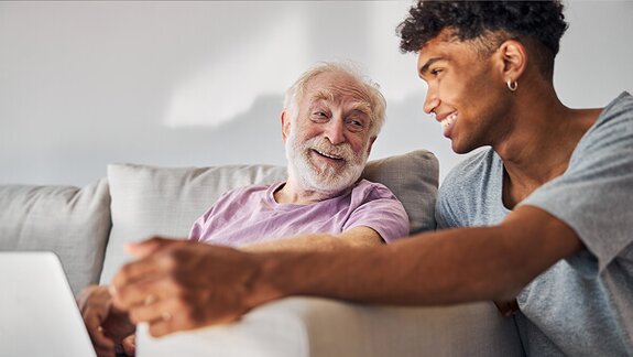 An older man and a younger man sit together on a couch talking while a laptop is open in front of them.
