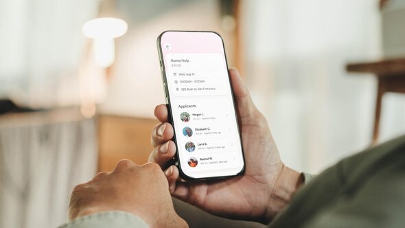 A person holds a smartphone displaying a list of applicants for a Home Help request, including their names, photos, distance, and application times.