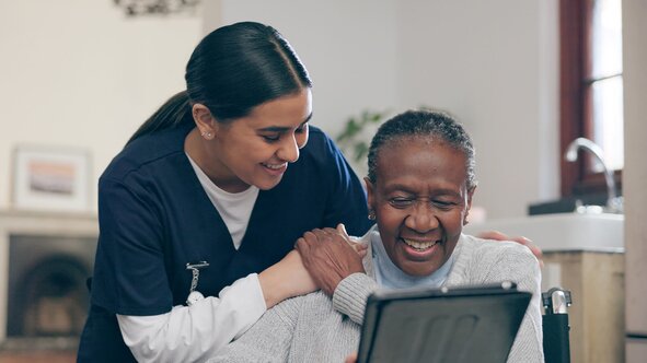 An older woman in glasses smiles up at her Helper, who stands behind her wheelchair indoors.