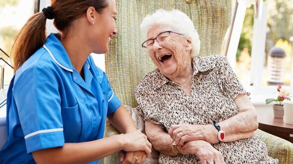 A Helper and a senior woman sit closely on a couch, smiling and laughing as they talk.